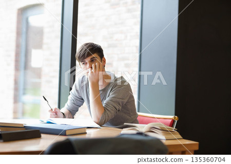 Male student sitting at wooden table by window holding pen over open notebook beside books 135360704