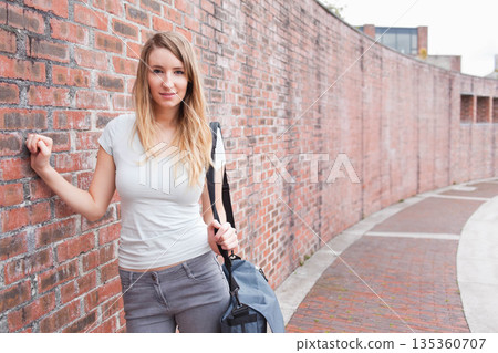 Woman leaning against curved red brick wall along paved walkway holding blue-gray duffel bag Woman leaning against curved red brick wall along paved walkway holding blue-gray duffel bag 135360707
