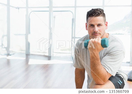 Man kneeling in fitness studio curling teal dumbbell at thigh next to glass wall, copy space 135360709
