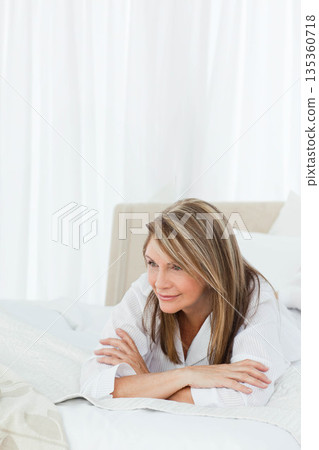 Senior woman reclining on white bed with beige headboard in bedroom gazing toward left, copy space 135360718