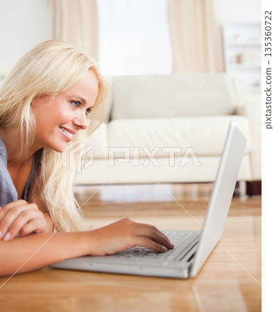 Blonde woman lying on floor typing on silver laptop beside light-colored sofa in living room 135360722