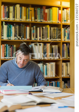 Senior man wearing gray sweater writing on notebook at wooden library table with textbooks and pen 135360727