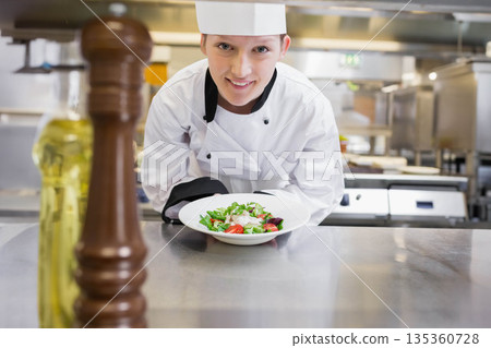 Female chef in uniform leaning at counter showing fresh salad with pepper mill in kitchen 135360728
