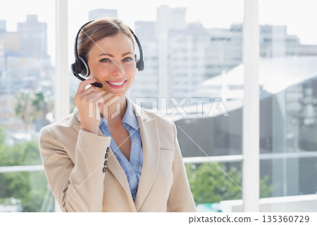 Mid adult woman standing in office wearing headset and smiling overlooking urban skyline via glass 135360729