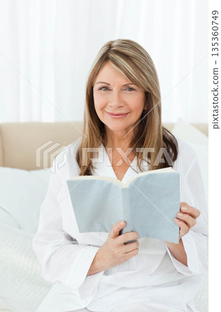 Senior woman reading light blue paperback book sitting on white bedding in bedroom with headboard Senior woman reading light blue paperback book sitting on white bedding in bedroom with headboard 135360749