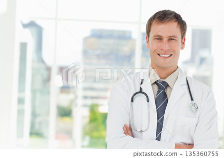 Mid adult male doctor standing arms crossed wearing lab coat and stethoscope in clinic, copy space Mid adult male doctor standing arms crossed wearing lab coat and stethoscope in clinic, copy space 135360755
