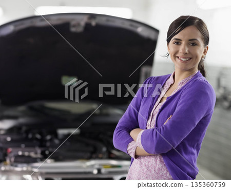 Woman standing with arms crossed in auto workshop beside open car hood and engine, copy space Woman standing with arms crossed in auto workshop beside open car hood and engine, copy space 135360759