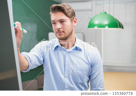 Man in his thirties wearing blue shirt marking presentation board with whiteboard marker in class Man in his thirties wearing blue shirt marking presentation board with whiteboard marker in class 135360778