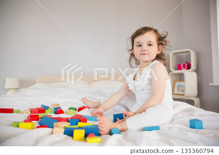 Toddler girl playing with colorful wooden blocks on white bed near pink storage boxes, copy space 135360794