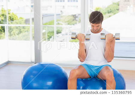 Man in sportswear sitting on stability ball holding dumbbells at shoulder level at gym, copy space Man in sportswear sitting on stability ball holding dumbbells at shoulder level at gym, copy space 135360795