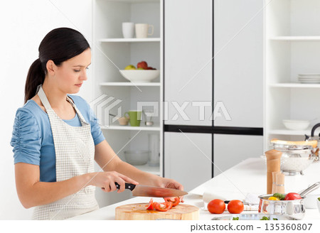 Woman chopping tomatoes on cutting board at kitchen counter wearing apron using knife, copy space 135360807