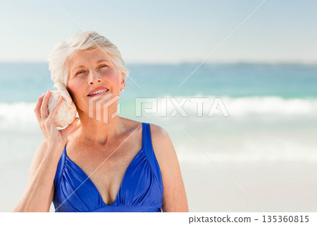 Senior female standing in blue swimsuit on sandy beach at shoreline holding conch seashell smiling 135360815