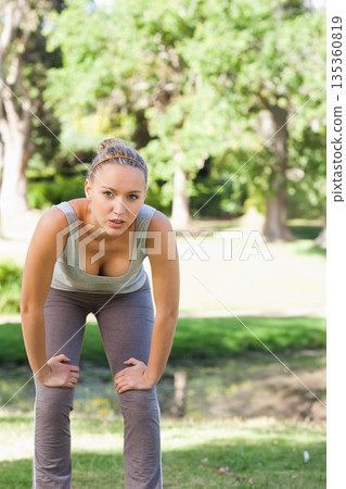 Female exerciser pausing workout by pond and catching breath in lush park wearing headband 135360819