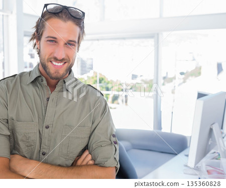 Man standing with arms crossed at office desk by window with computer monitor, copy space 135360828