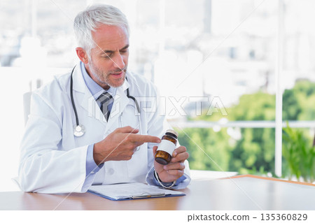 Senior male doctor holding clipboard pointing at amber pill bottle wearing stethoscope at desk Senior male doctor holding clipboard pointing at amber pill bottle wearing stethoscope at desk 135360829