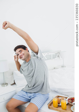 Male wearing grey tee and checkered shorts stretching arms overhead on bed beside tray with juice Male wearing grey tee and checkered shorts stretching arms overhead on bed beside tray with juice 135360830