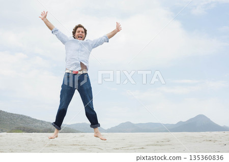 Man jumping barefoot wearing light shirt and rolled jeans on sandy beach under clouds Man jumping barefoot wearing light shirt and rolled jeans on sandy beach under clouds 135360836