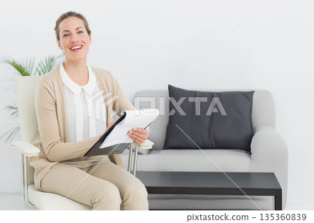 Mid adult woman sitting on white swivel chair in bright consultation room holding clipboard and pen Mid adult woman sitting on white swivel chair in bright consultation room holding clipboard and pen 135360839
