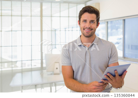 Man in grey polo shirt holding tablet beside desk and glass partition at office, copy space 135360844
