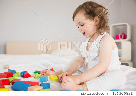 Toddler girl playing with colorful wooden blocks on bed in room wearing amber necklace, copy space Toddler girl playing with colorful wooden blocks on bed in room wearing amber necklace, copy space 135360851