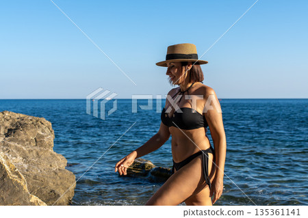 Woman beach ocean, confident female wearing bikini and hat walking along rocky coast enjoying summer vacation with copy space Woman beach ocean, confident female wearing bikini and hat walking along rocky coast enjoying summer vacation with copy space 135361141