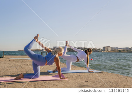 Yoga, Beach, Women - Two women practice yoga on a mat by the sea on a sunny day. Yoga, Beach, Women - Two women practice yoga on a mat by the sea on a sunny day. 135361145