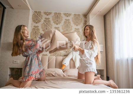 Pillow Fight Bedroom Fun: Two Women playfully fighting pillows on a bed in a modern bedroom, enjoying leisure time. 135361236