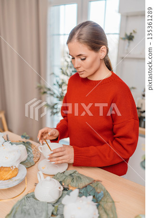 Young woman stirring tea in cup preparing breakfast in cozy bright kitchen 135361289