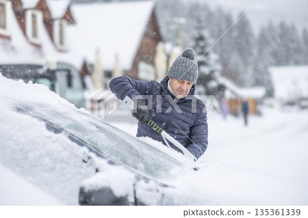 A man in a dark jacket and gray hat diligently removes a thick layer of fresh snow from the windshield of his car with a scraper and a broom. The entire scene takes place during a heavy snowfall in a 135361339