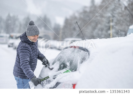 A man in a dark jacket and gray hat diligently removes a thick layer of fresh snow from the windshield of his car with a scraper and a broom. The entire scene takes place during a heavy snowfall in a 135361343