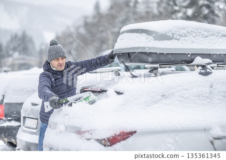A man in a blue jacket and knit hat uses a broom to clear a thick layer of snow from the back of his car. The surrounding snowy landscape and the roof box on the vehicle suggest that he is preparing A man in a blue jacket and knit hat uses a broom to clear a thick layer of snow from the back of his car. The surrounding snowy landscape and the roof box on the vehicle suggest that he is preparing 135361345