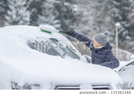 A man in a dark jacket and gray hat diligently removes a thick layer of fresh snow from the windshield of his car with a scraper and a broom. The entire scene takes place during a heavy snowfall in a 135361348
