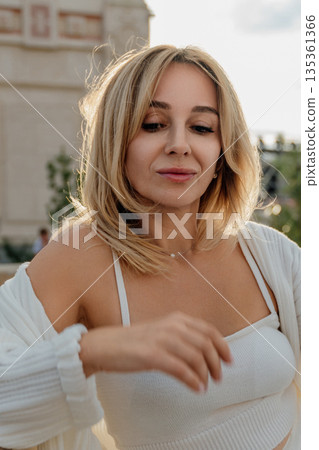 Woman Blonde Hair Outdoor Casual - A young woman with blonde hair stands outdoors in a casual white outfit. She has a relaxed expression and is looking down. Woman Blonde Hair Outdoor Casual - A young woman with blonde hair stands outdoors in a casual white outfit. She has a relaxed expression and is looking down. 135361366