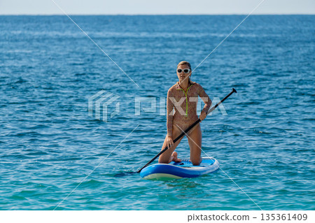 Woman, paddleboard, ocean. Active female kneeling on a stand up paddleboard in clear blue water enjoying summer sports with copy space. 135361409