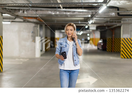 A woman in a denim jacket walks through a parking garage while talking on her phone and holding a coffee cup A woman in a denim jacket walks through a parking garage while talking on her phone and holding a coffee cup 135362274