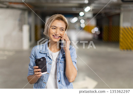 A smiling woman in a parking garage holds a coffee cup while talking on her phone A smiling woman in a parking garage holds a coffee cup while talking on her phone 135362297