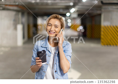 A smiling woman in a denim jacket talks on her phone while holding a coffee cup in a parking garage 135362298
