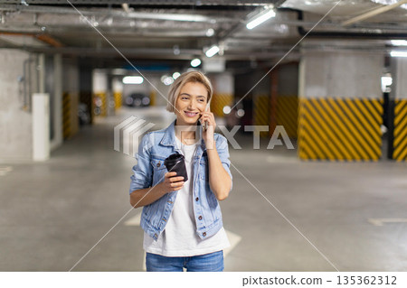 A woman smiles while talking on her phone and holding a coffee cup in a parking garage A woman smiles while talking on her phone and holding a coffee cup in a parking garage 135362312