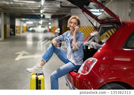 A woman smiles while talking on her phone, sitting in the open trunk of a red car in a parking garage A woman smiles while talking on her phone, sitting in the open trunk of a red car in a parking garage 135362313