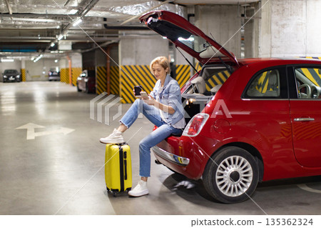 A woman sits in the open trunk of a red car in an underground parking garage, using her phone and enjoying a coffee A woman sits in the open trunk of a red car in an underground parking garage, using her phone and enjoying a coffee 135362324