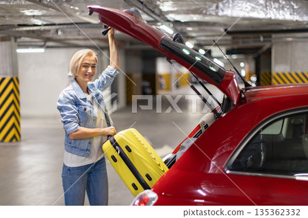 A smiling woman loads a yellow suitcase into the trunk of a red car in a parking garage A smiling woman loads a yellow suitcase into the trunk of a red car in a parking garage 135362332
