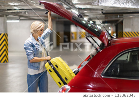 A woman smiles while loading a yellow suitcase into the trunk of a red car in a parking garage A woman smiles while loading a yellow suitcase into the trunk of a red car in a parking garage 135362343