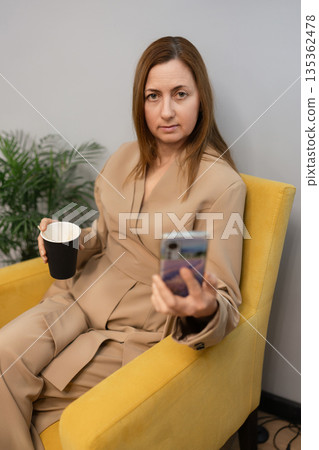 Businesswoman smartphone relaxing coffee break in yellow armchair corporate lounge Businesswoman smartphone relaxing coffee break in yellow armchair corporate lounge 135362478