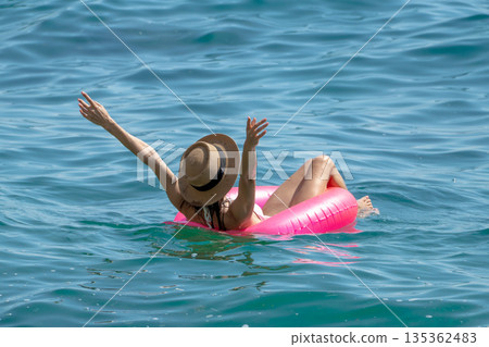 Woman Floating Sea: Woman relaxing on pink inflatable in blue sea, hat on, arms raised, summer fun. Woman Floating Sea: Woman relaxing on pink inflatable in blue sea, hat on, arms raised, summer fun. 135362483