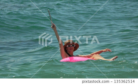 Woman Tablet Ocean. Woman floats on pink ring, raising tablet to capture sunny sea view on summer vacation. 135362514