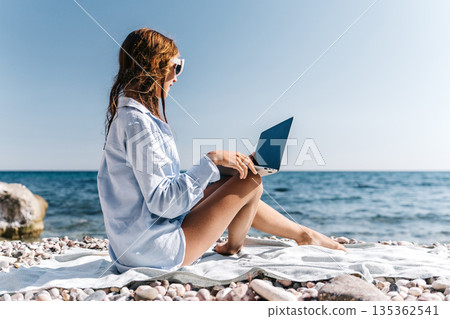 Woman, laptop, beach. Young woman uses computer by the ocean on a sunny pebble shore, remote work concept, copy space Woman, laptop, beach. Young woman uses computer by the ocean on a sunny pebble shore, remote work concept, copy space 135362541
