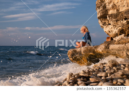 Child, Coast, Sea - Young boy sits on cliff overlooking the ocean with waves crashing on the rocks. 135362557