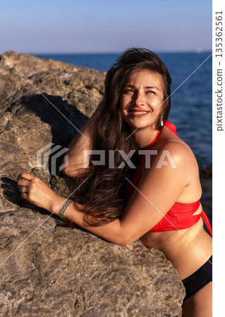 Woman, beach, rocks. Happy young woman smiling, leaning on sunlit rocks by the ocean with copy space. Woman, beach, rocks. Happy young woman smiling, leaning on sunlit rocks by the ocean with copy space. 135362561