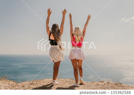 Two women are standing on a beach, one wearing a pink top and the other wearing a black top. They are both smiling and raising their arms in the air, as if they are celebrating something. Two women are standing on a beach, one wearing a pink top and the other wearing a black top. They are both smiling and raising their arms in the air, as if they are celebrating something. 135362588