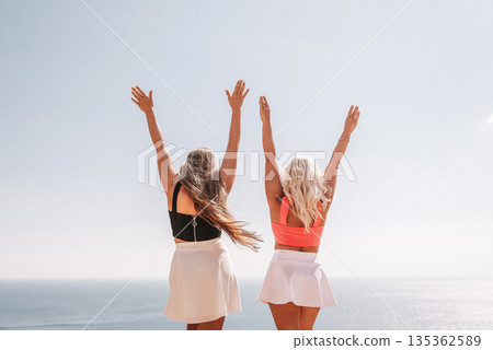 Two women are standing on a beach, one wearing a pink top and the other wearing a black top. They are both smiling and raising their arms in the air, as if they are celebrating something. 135362589